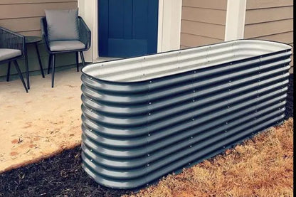 A modular raised garden bed in dark gray, made from metal, situated on grass with a building and chair in the background.